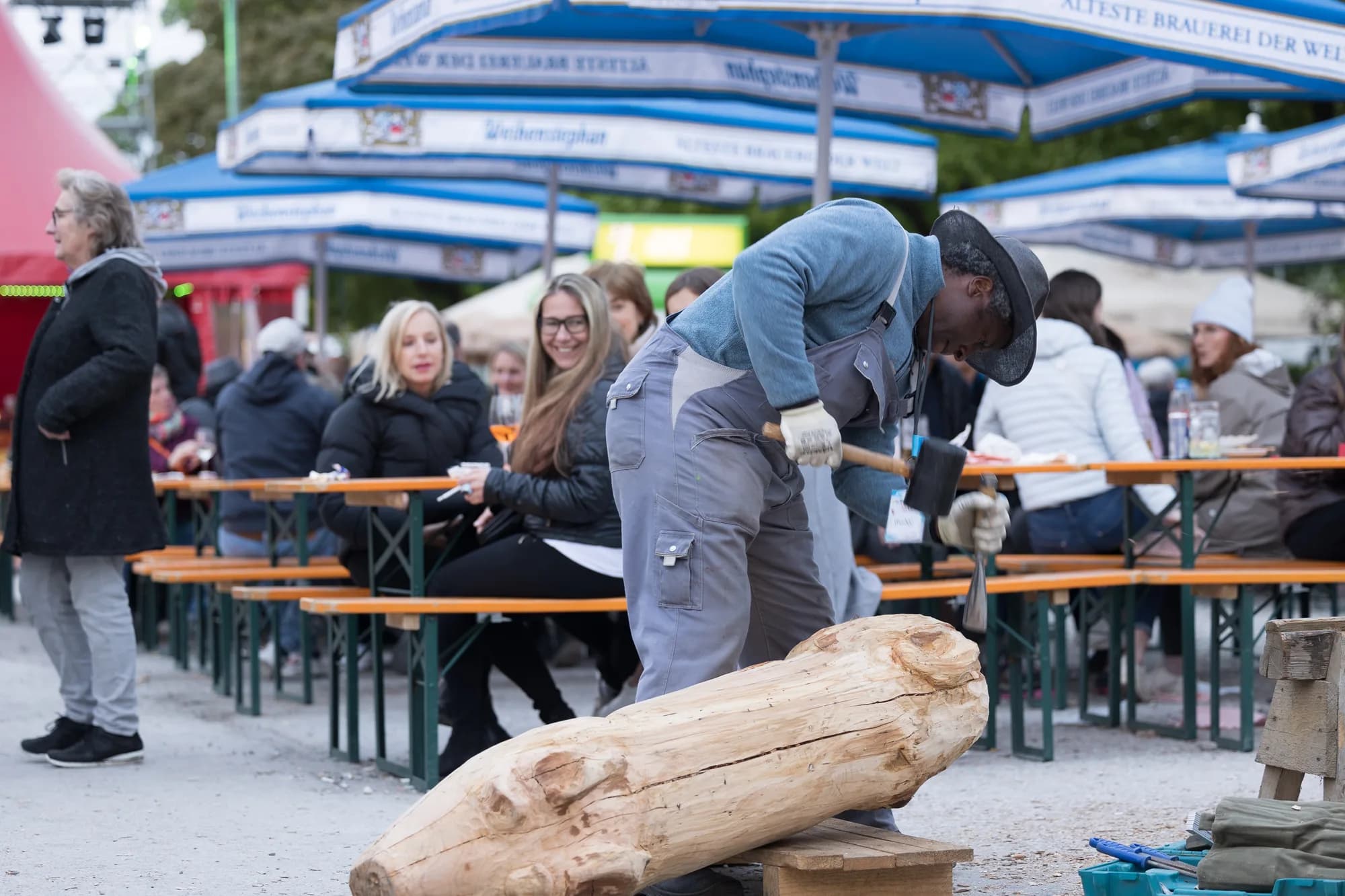 A woodcarver in blue overalls and a hat uses a mallet and chisel on a large log in front of seated festival guests.