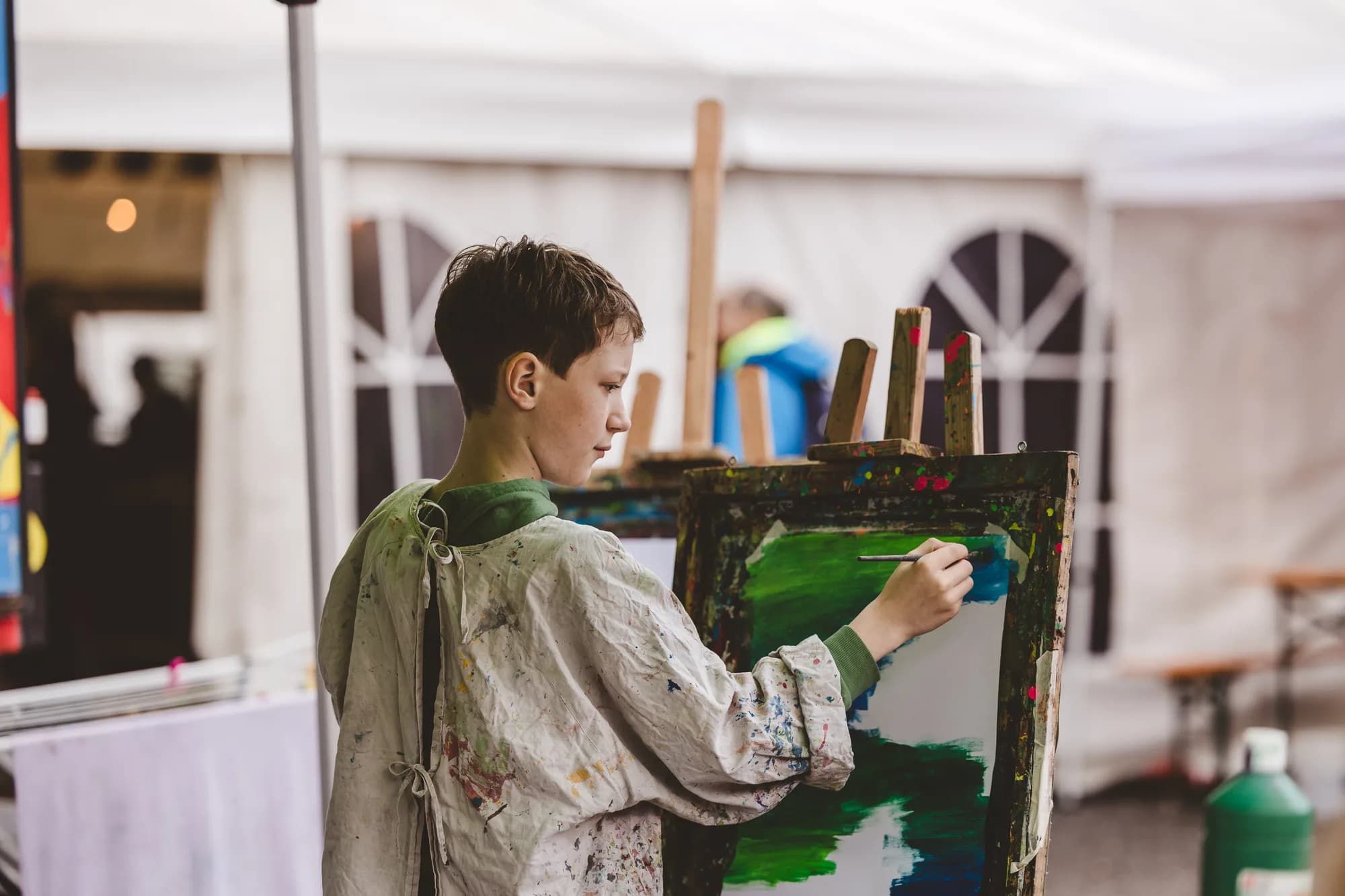 A young boy in a paint-splattered smock concentrates on painting a green landscape on a canvas inside a white festival tent.